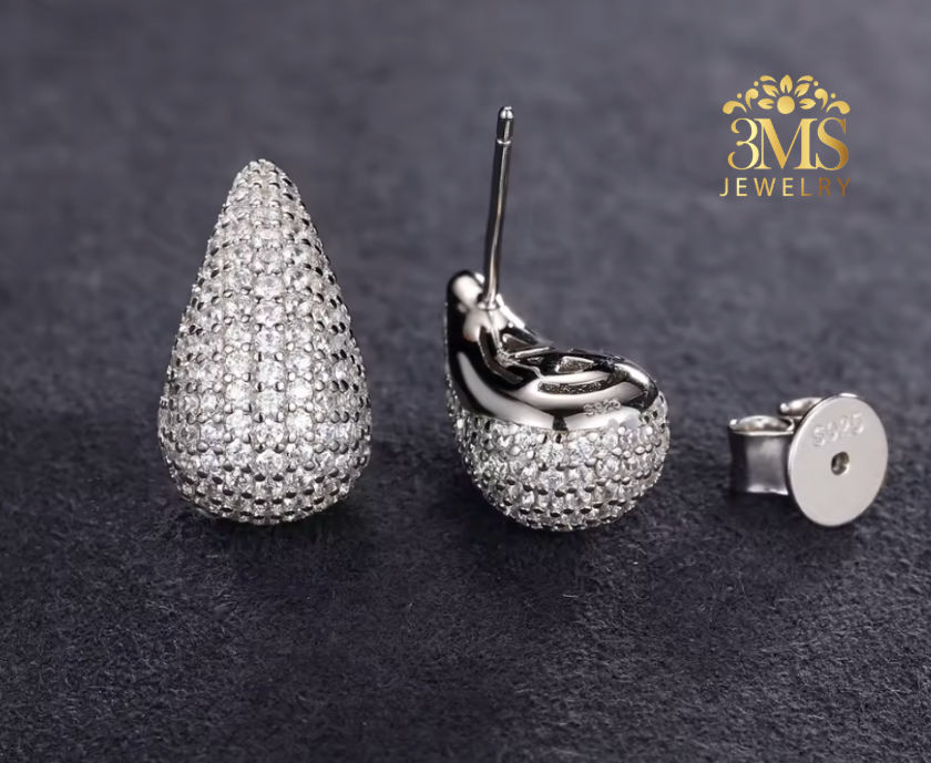 Teardrop-shaped silver earrings with clear stones on a dark background, branded '3MS Jewel By'.