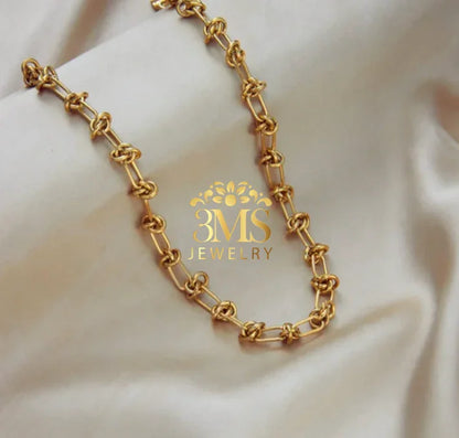 Gold chain necklace with unique knot links displayed on soft beige fabric background