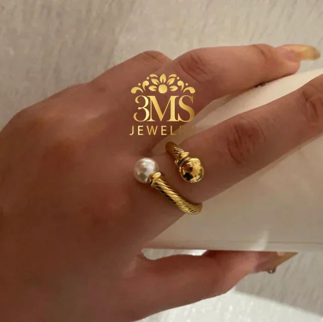 Gold open ring with twisted design featuring a pearl on one end and a golden ball on the other worn on a hand