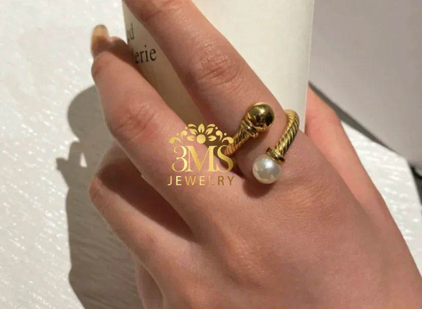 Close-up of hand wearing gold twisted ring with pearl and gold bead against white textured background